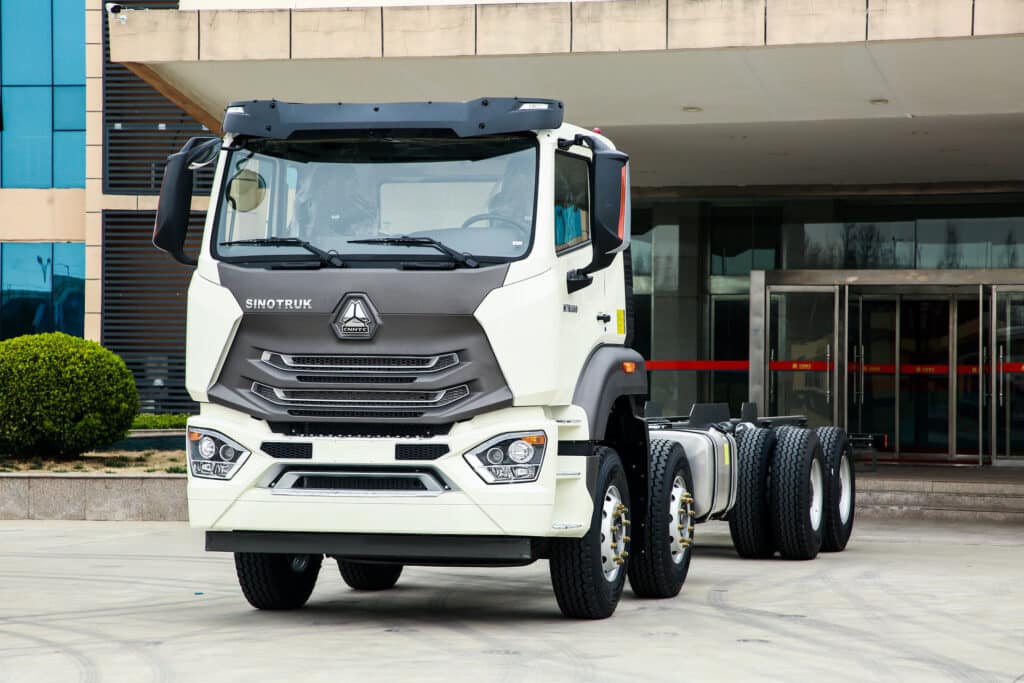 A white Sinotruk heavy-duty truck cab with six wheels is parked outside a modern building with glass doors and windows.